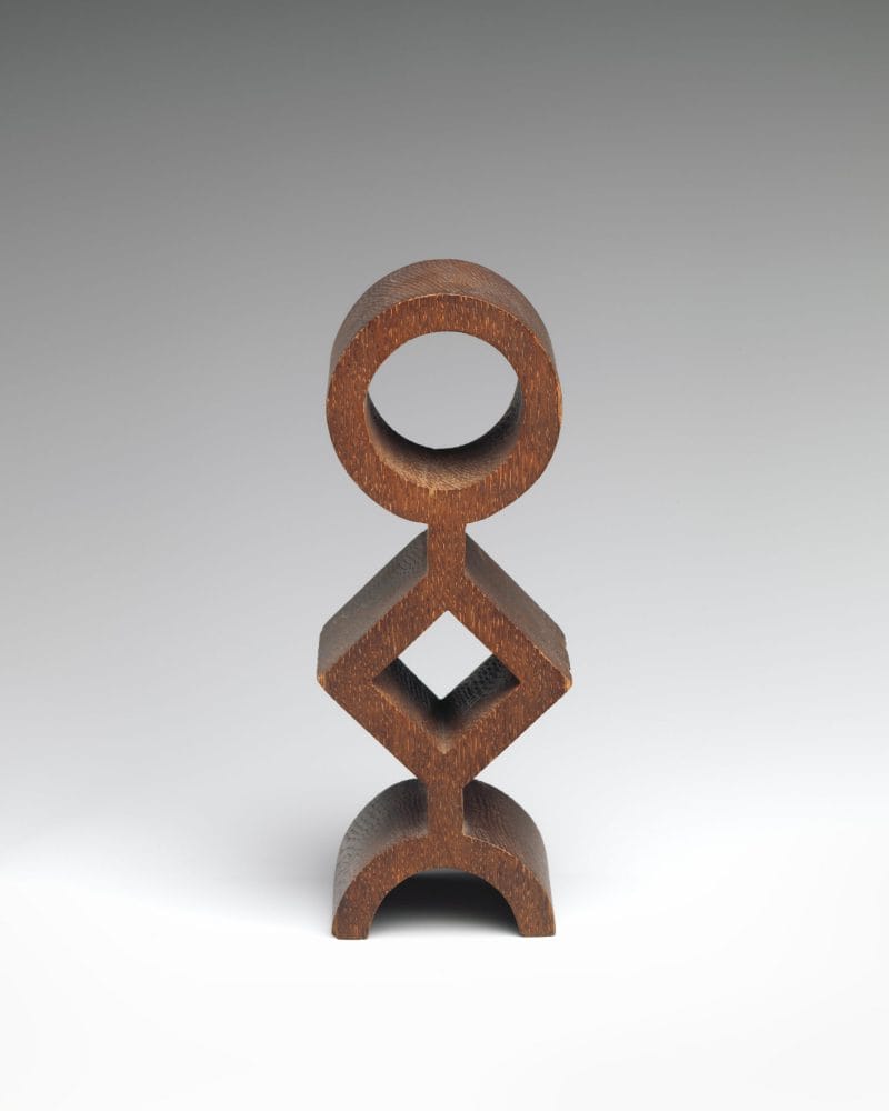 A wooden totem-like sculpture composed of a hollow circle stacked on a hollow diamond stacked on a semi-circle.