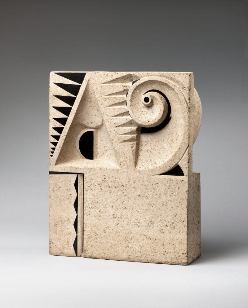 Rectangular limestone block carved with abstractions of geometric shapes, inlay of black stone in triangular patterns, and a spiral that loops at the top-right corner.