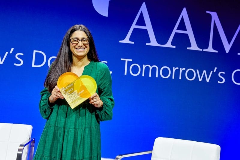 Mona Hanna-Attisha, holding a heart-shaped award, stands on a stage with a blue backdrop of the AAMC.