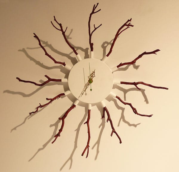 An off-white wall clock with dark red twigs depicting the hours with two gold ornate clock hands.
