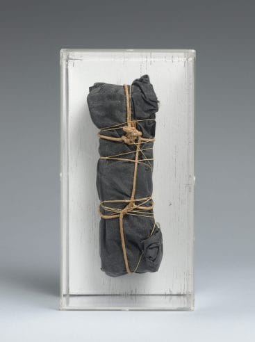 A package wrapped in black fabric with twine and rope and attached to a white block of wood and enclosed in plexiglass.