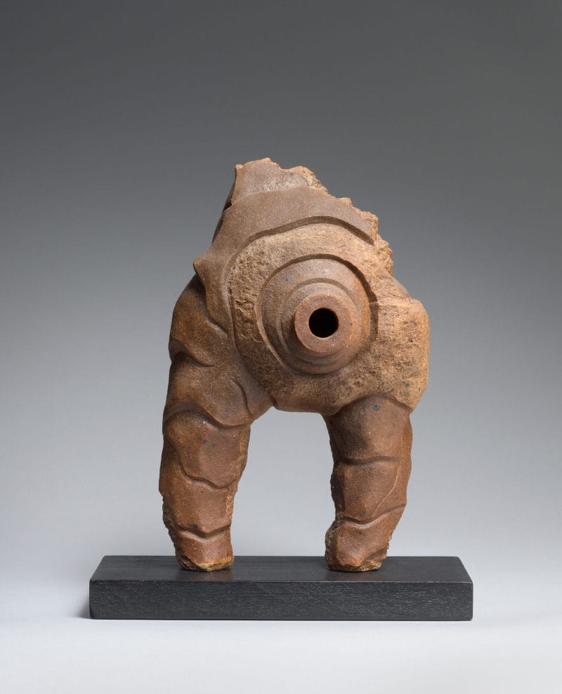 Amorphous brown sculpture with a centered hole, standing upon a short dark gray rectangular pedestal.