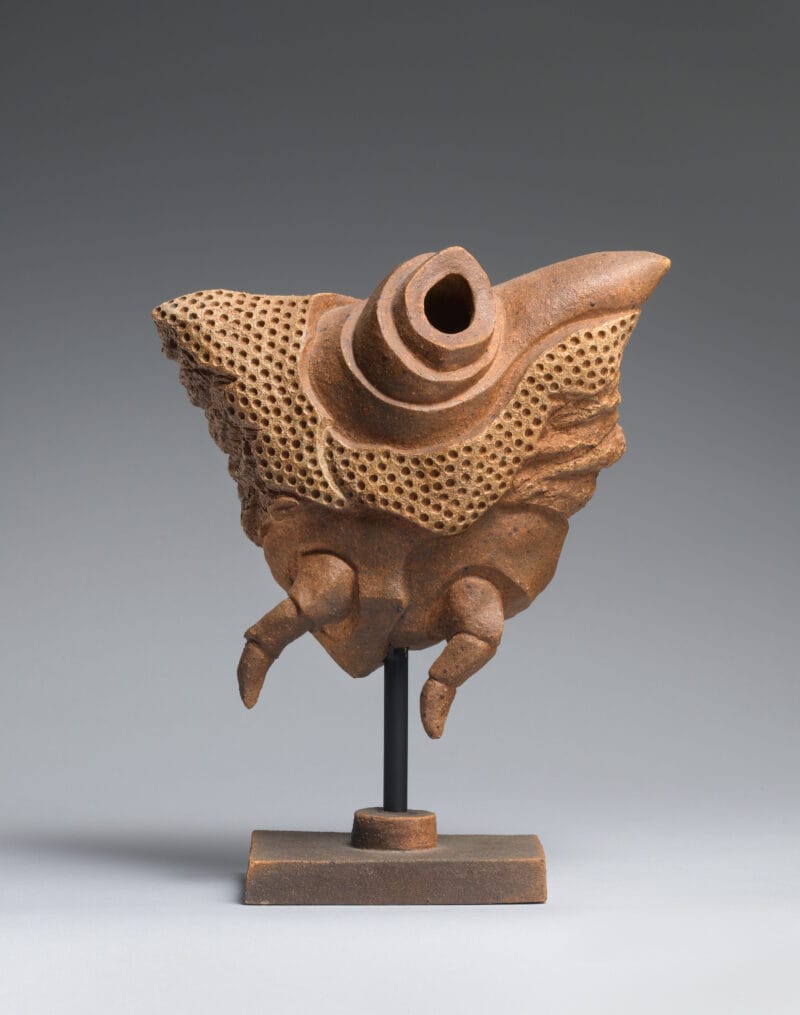 Brown triangular sculpture with mulitple small holes, two smalls legs, and a large hole atop, held up on a short brown pedestal.