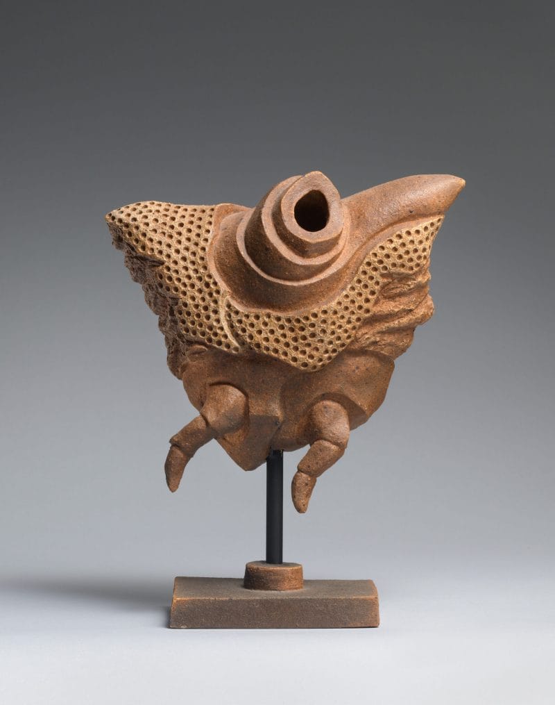 Brown triangular sculpture with mulitple small holes, two smalls legs, and a large hole atop, held up on a short brown pedestal.