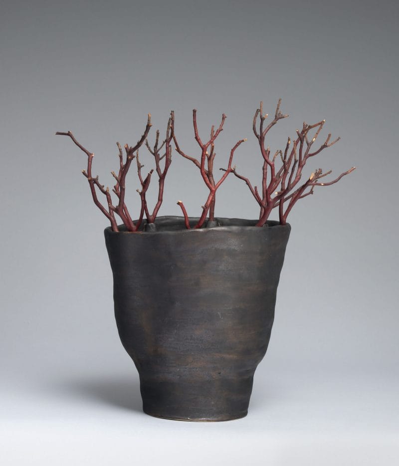 A black vase with an uneven surface holding dark red twigs.