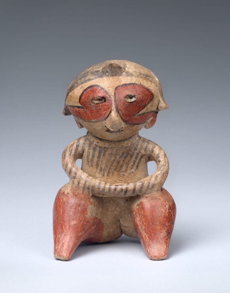 Seated ceramic figure with hands rested on knees, and face and body tattoos in red and black ink.