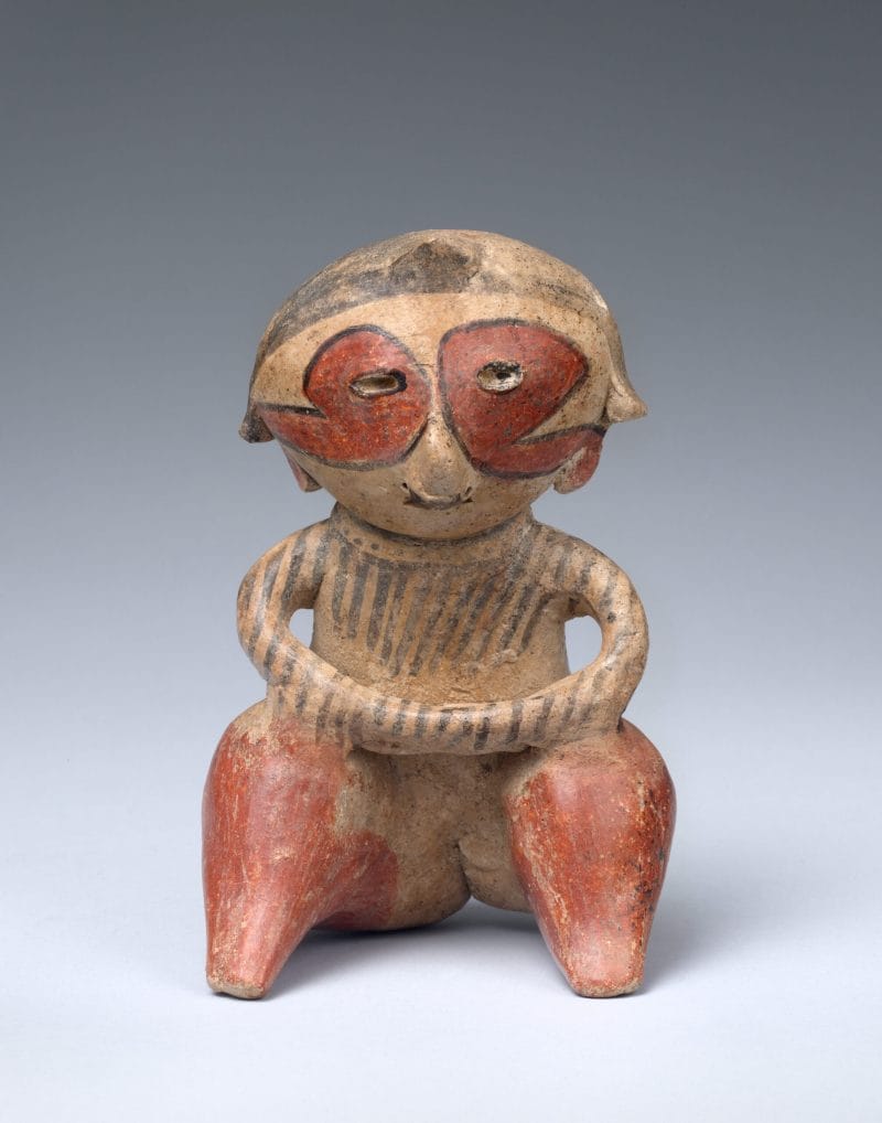 Seated ceramic figure with hands rested on knees, and face and body tattoos in red and black ink.