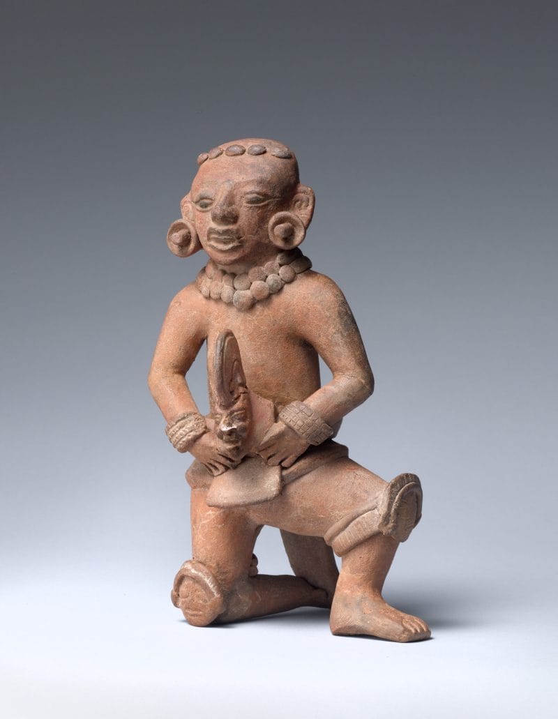 Ceramic sculpture of a ballplayer kneeling on one knee wearing knee pads, hand mitts, and waist pads, and earrings while holding a head.