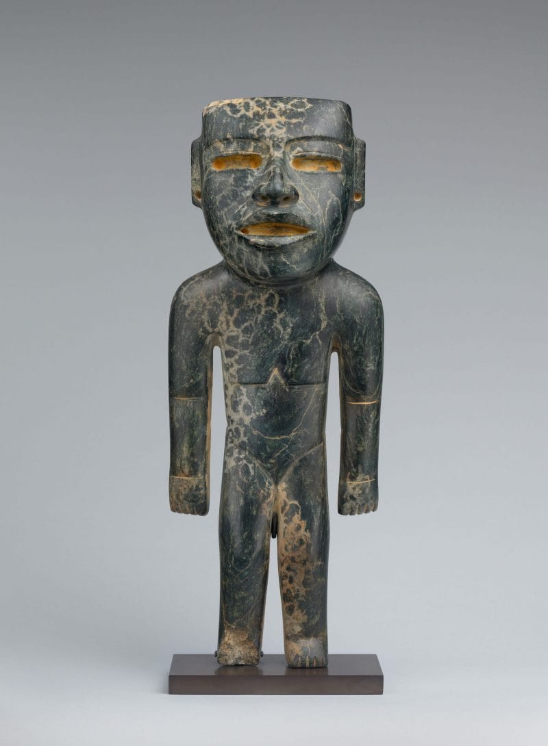 Standing stone figure with a face resembling an Olmec mask with deeply cut eyes and mouth.