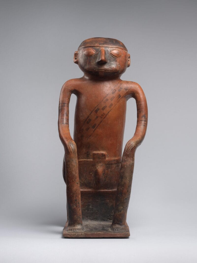 A seated male figure with a painted face, enlarged cheeks, and a woven band across the chest.