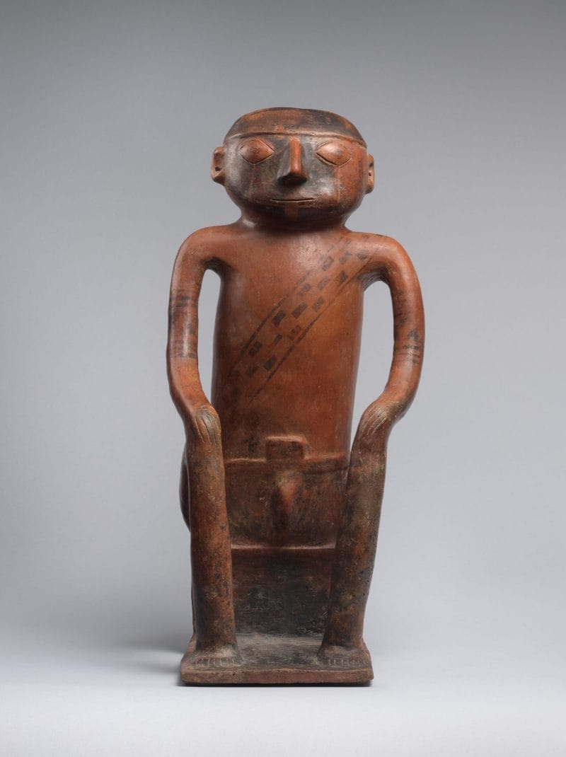A seated male figure with a painted face, enlarged cheeks, and a woven band across the chest.