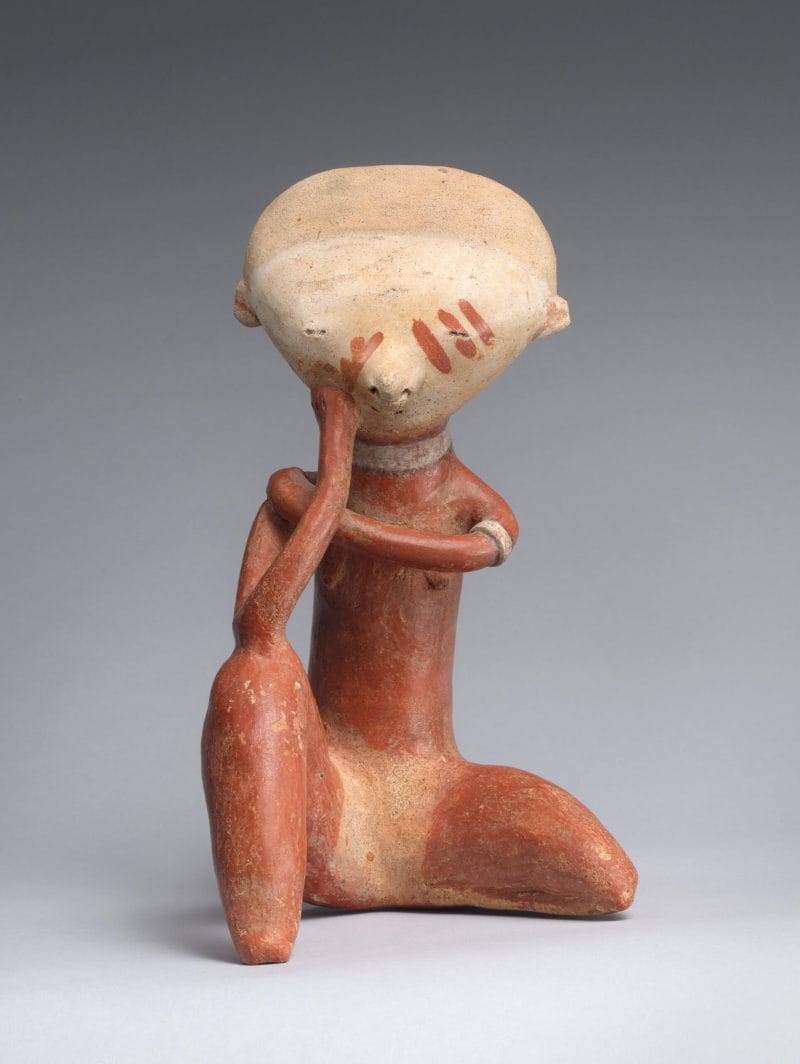 A stylized ceramic figure with a red body and beige head, red face paint, and a hand resting on its face.