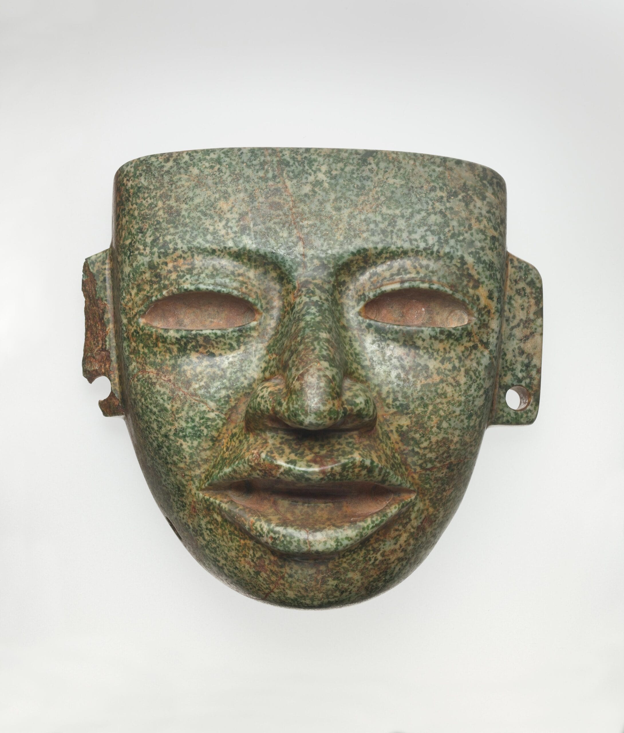 Mottled green stone face mask with open mouth, hollowed eyes, and pierced ears.
