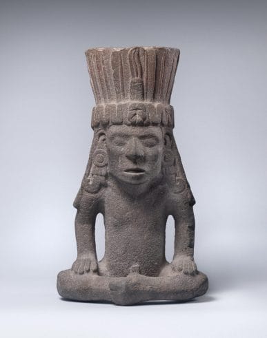 Sculpture of a man seated with crossed legs and wearing a large headdress and long, intricate earings; his mouth is open as if he is in mid-speech.
