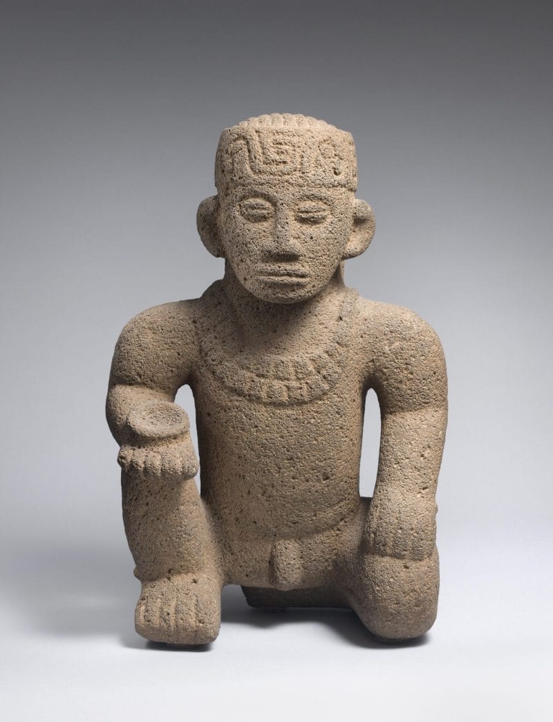 Stone sculpture of man kneeling on one knee, holding a small dish, wearing a beaded necklace and headdress.