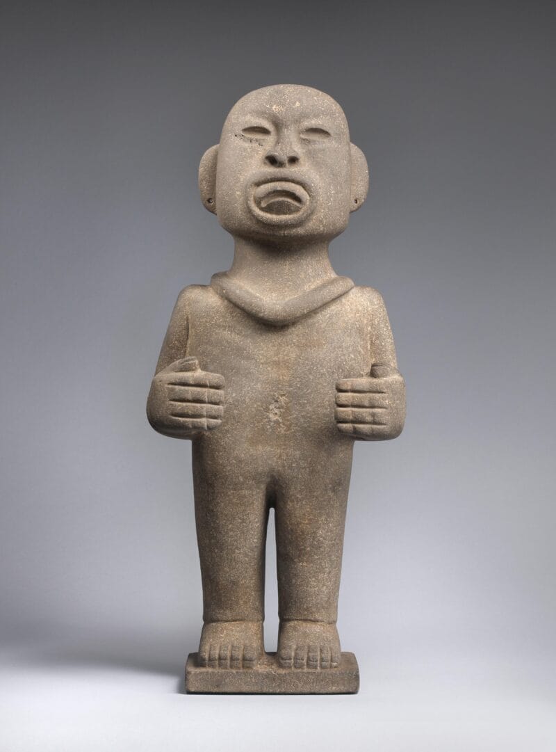 Gray standing stone figure with indented eyes and mouth, pierced ears, a bald head, and bent arms.