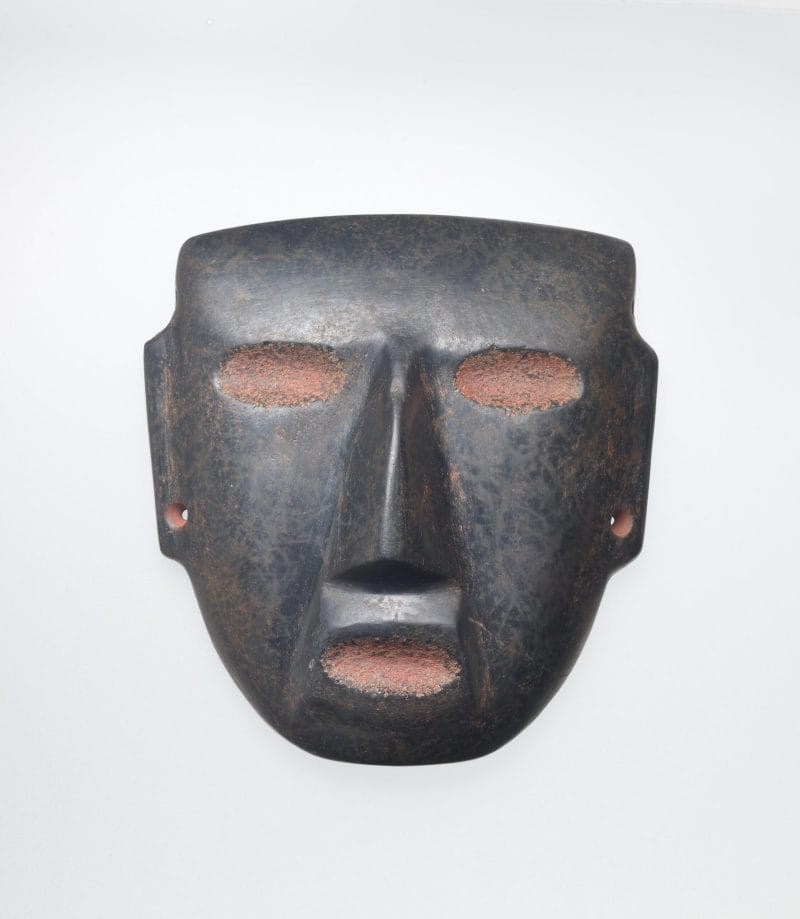 Black stone mask with red eyes and mouth, drilled ear spools, and a triangular nose.