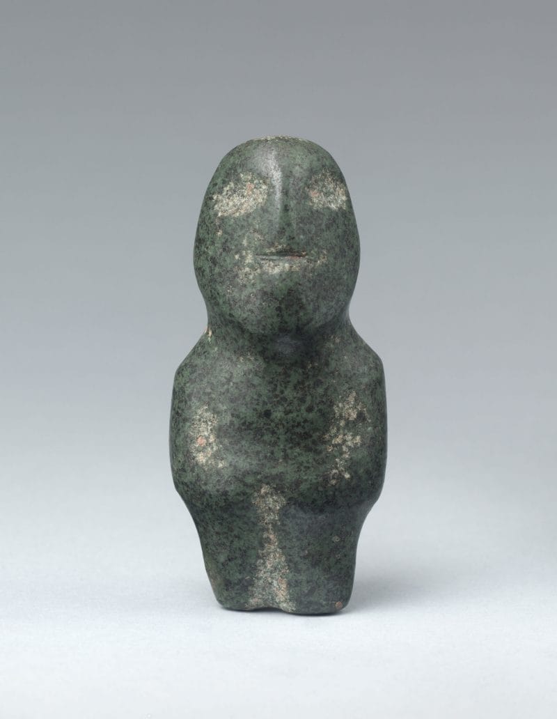 A standing green stone figure with arms crossed and small, hollow facial features.
