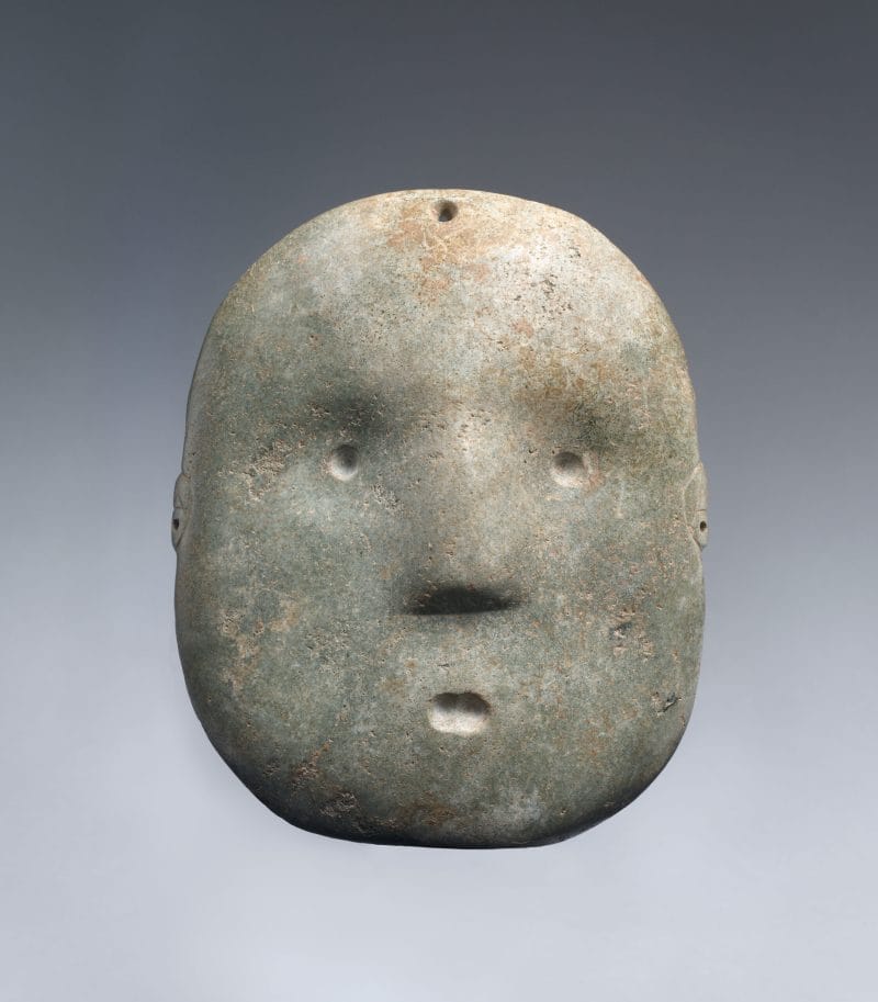 A light green stone face mask with rounded form, sharp nose, and simplified features.