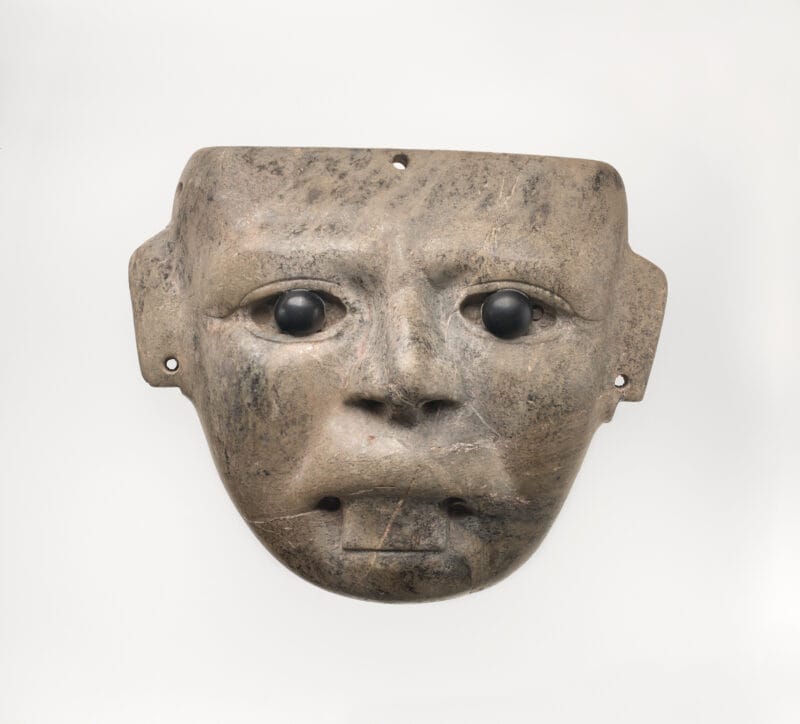 Stylistic stone mask with inlaid black eyes and stuck out tongue.