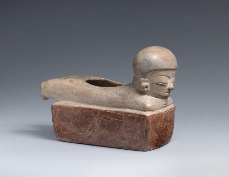 Ceramic vessel of a human figure with a helmet and earplugs, laying on their stomach.