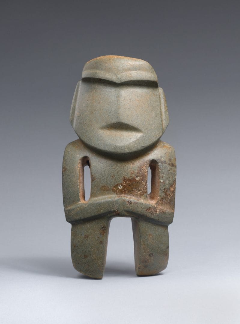 Stone sculpture of an abstracted standing figure with carved facial features, and arms resting across the torso.