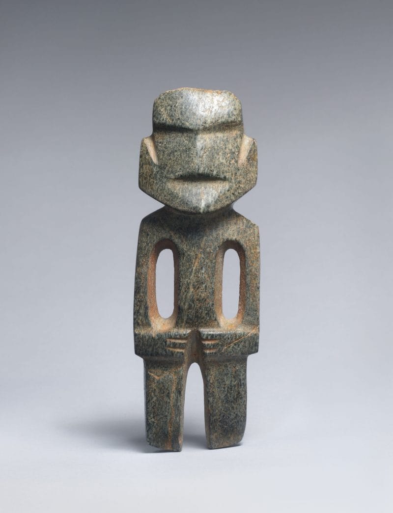 Stone figure with a broad rounded face, indentations for the nose and mouth, two hollows to form the torso and arms, and hands folded across the body.