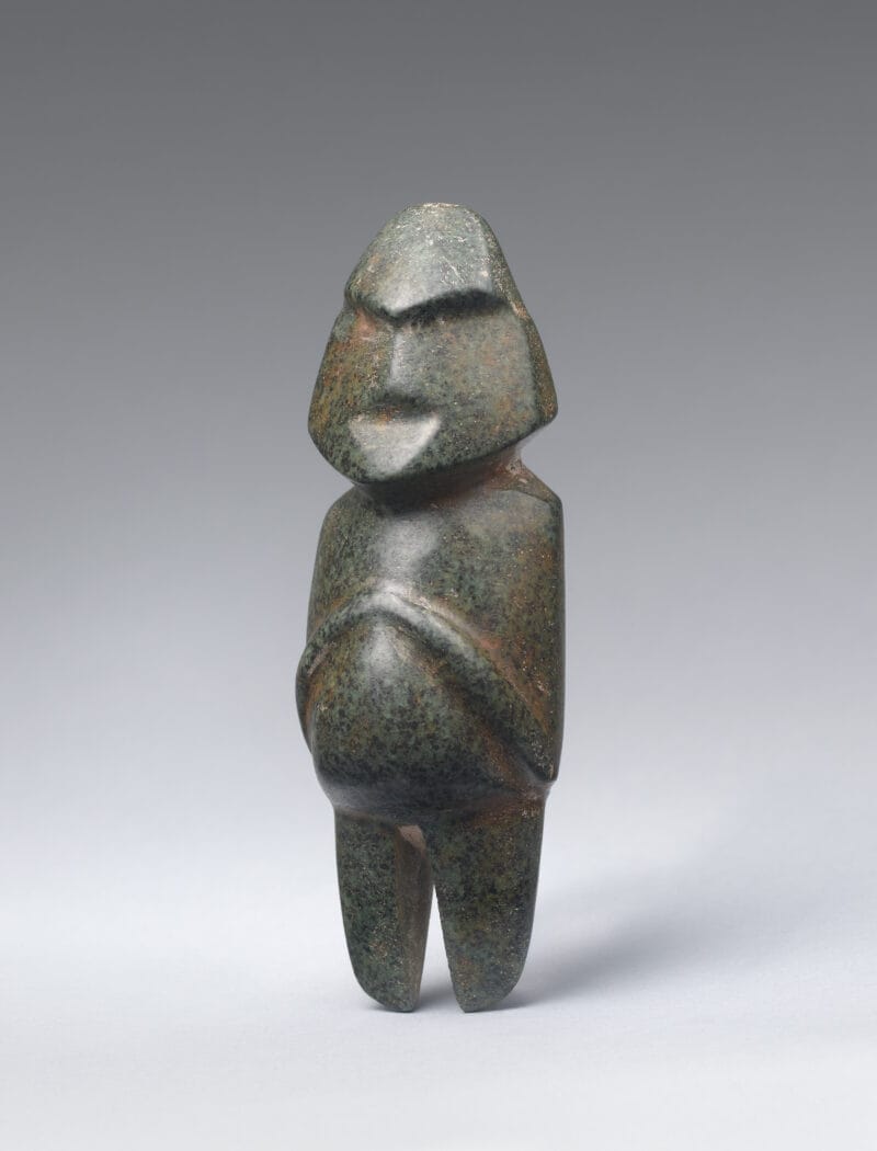 Small, abstract stone carving of a standing human figure with broad, pointed facial features and folded hands upon its round abdomen.