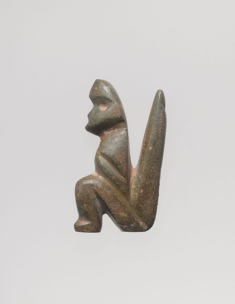 Stone figure seated in profile with verticle cuts to define physical features, and a long rigid tail behind it.