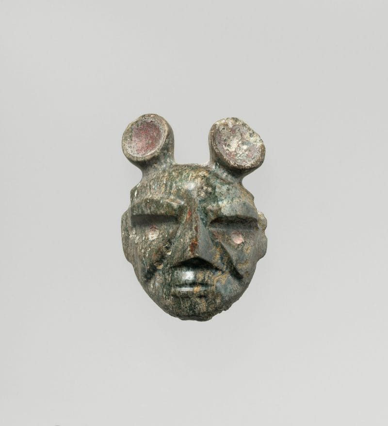 Two protruding tube-like ears atop a human head carved from stone with abstract facial features defined by vertical cuts.