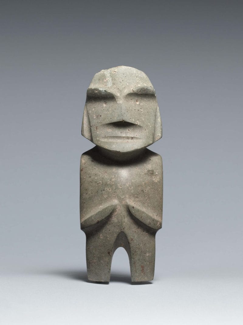 Abstract standing figure with indented facial features and pointed nose, arms at the side and folded at the ribs.