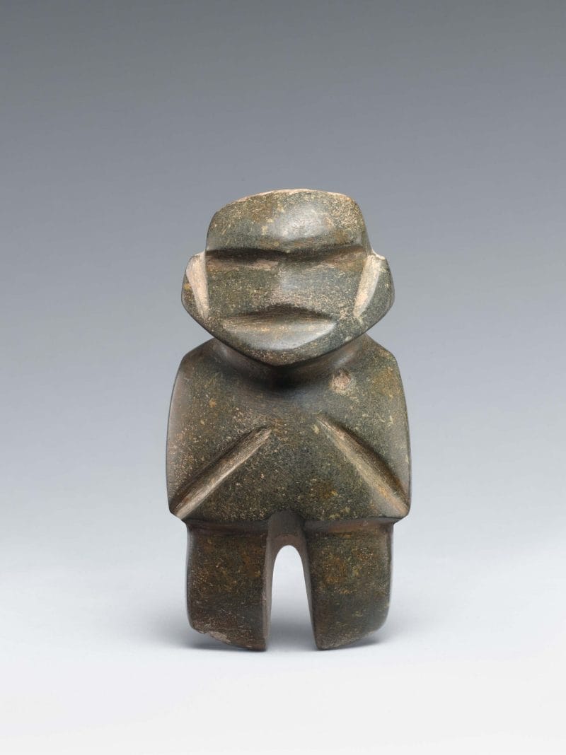 Abstract dark-green standing stone figure with indented features, including eyes, ears, mouth, arms, and legs.