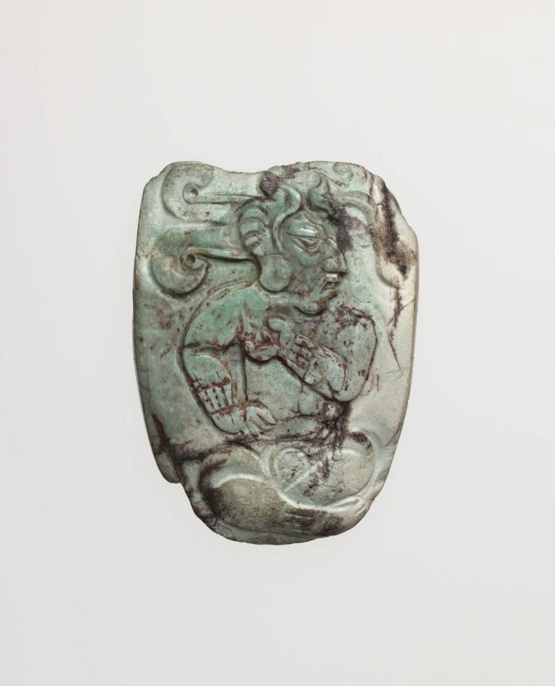Light green stone pendant with a carving of a dignitary shown in profile, seated cross-legged, his body facing forward, wearing an elaborate headdress.