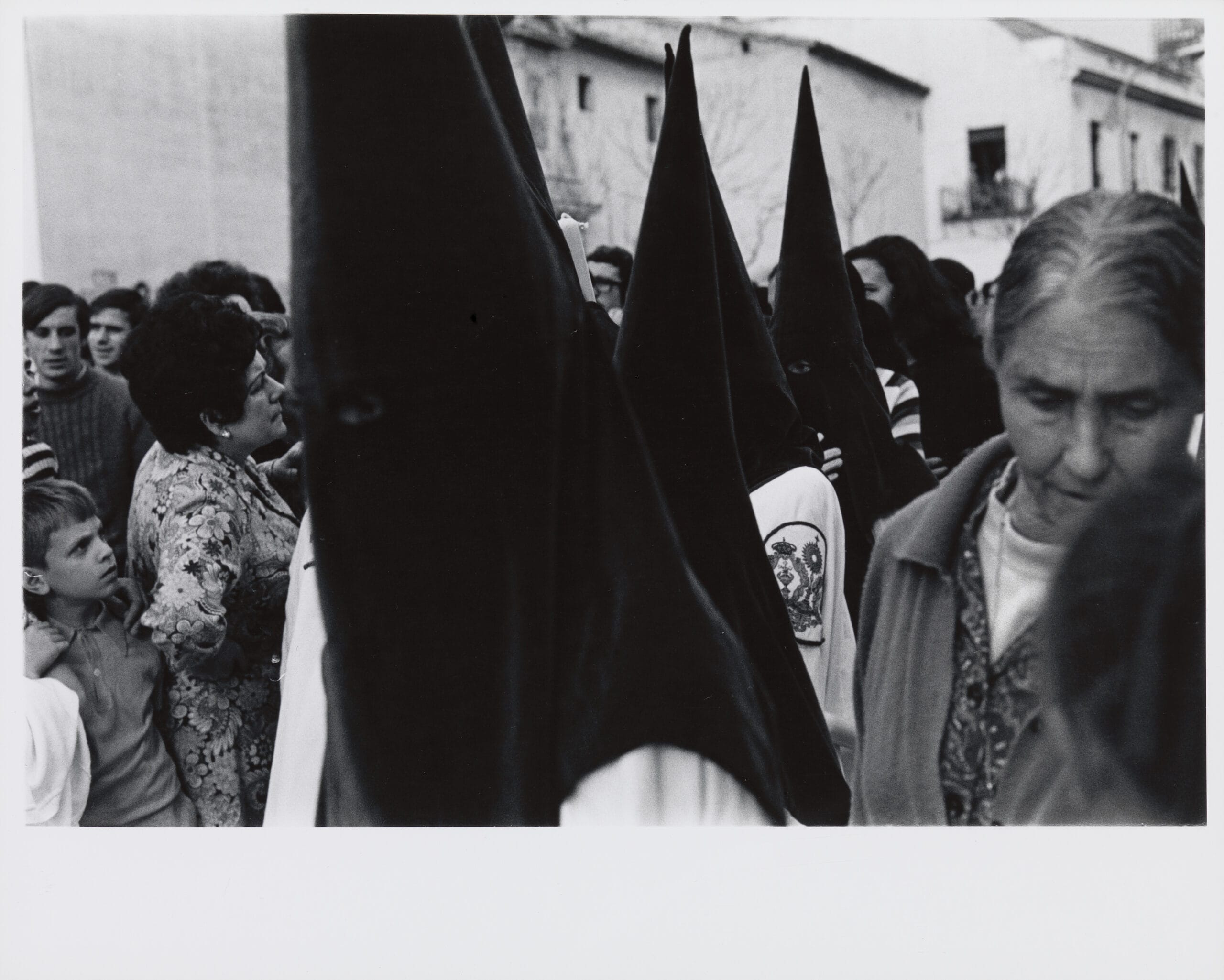 A black and white photograph of a procession of hooded penitents in Seville, Spain during Holy Week.