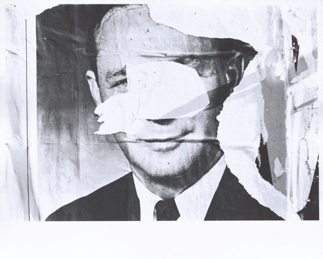 Black and white photograph of layered and torn signs that include the face of a man that has been torn leaving only a fragment of his face.