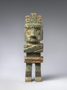 Standing stone figure with large goggle-like eyes, a curled lip, large teeth, folded arms wearing a headdress.