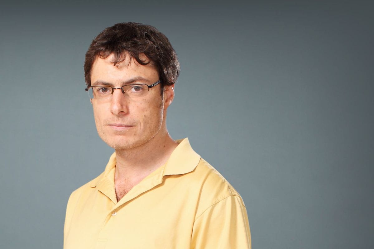 Evgeny Nudler, wearing a yellow polo shirt, in front of a neutral gray background.