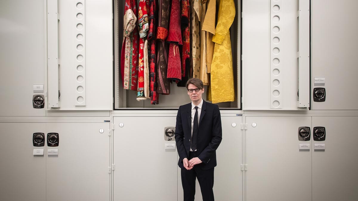 Andrew Bolton stands in front of a locker containing garments made of red and yellow silk, wearing a black suit and tie.