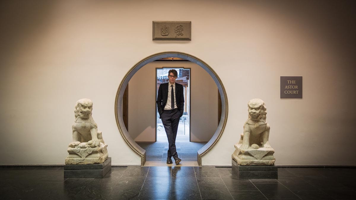 Andrew Bolton, dressed in a suit, stands in the round entry way of the Astor Court, with two lion statues on each side.