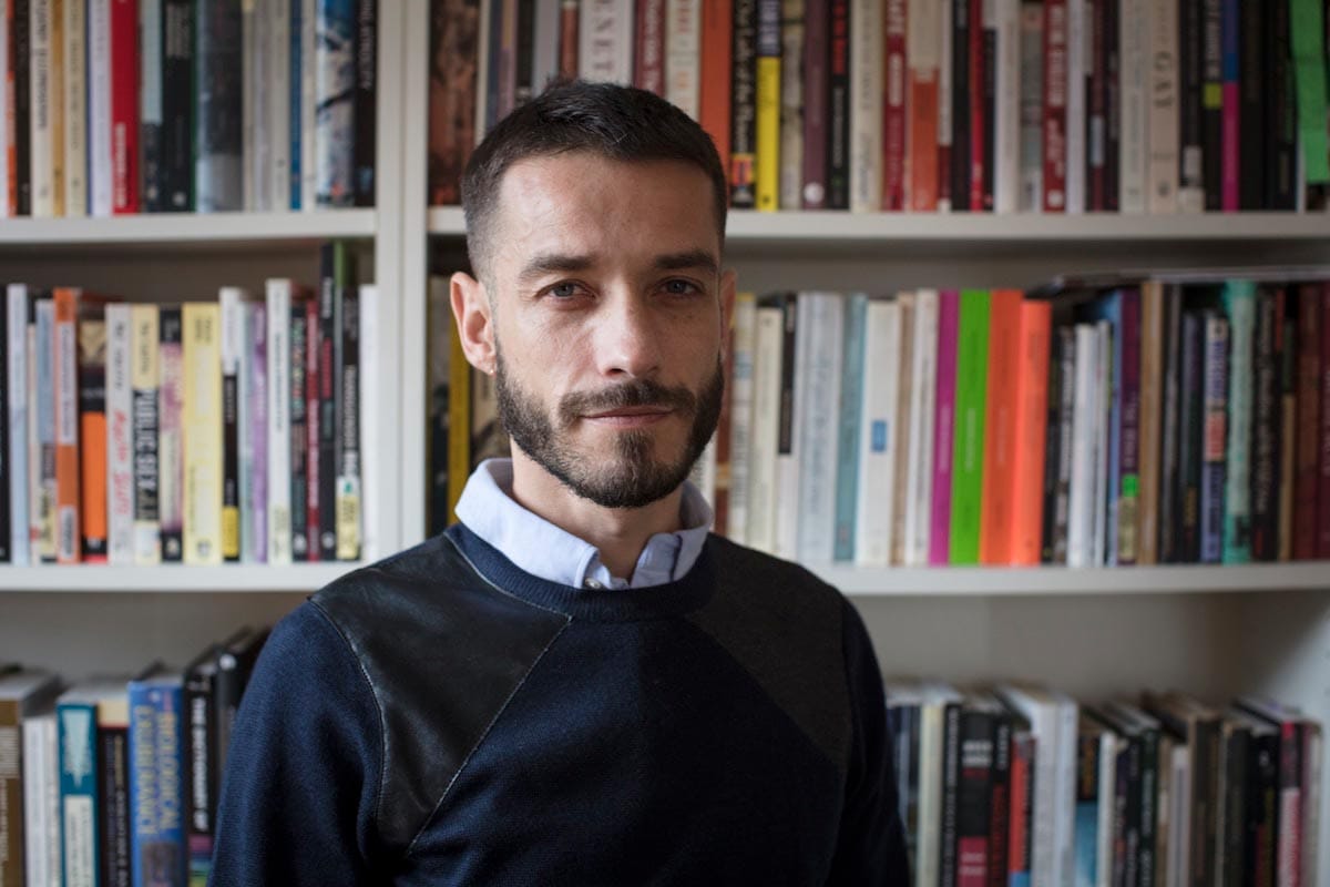 Carlos Motta is pictured from the chest-up in front of a bookshelf full of books.