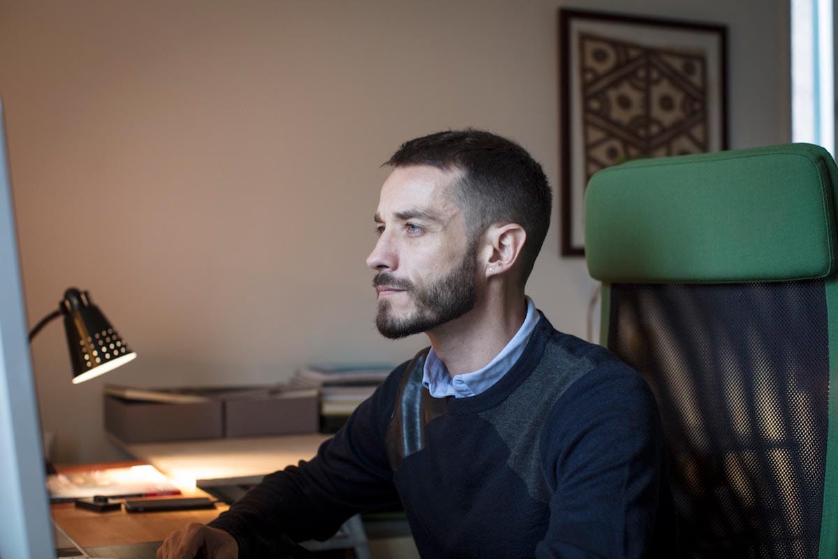 Carlos Motta in profile is pictured chest-up sitting in front of a computer screen.