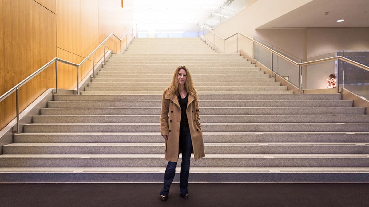 Franziska Michor stands at the foot of a large, wide staircase at a research facility, dressed in a trench coat.