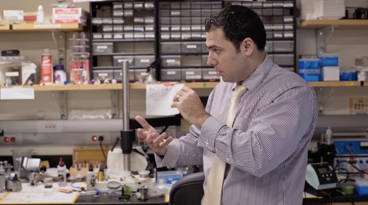 Michael Halassa stands in front of shelves filled with lab apparatus, actively conversing with his researchers (not pictured.)