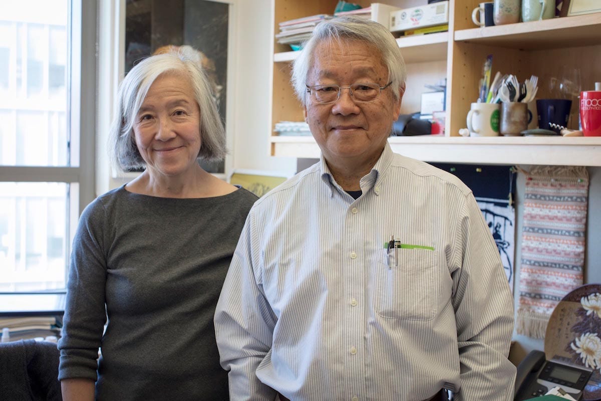 Pictured from the waist-up, Lily and Yuh-Nung Jan stand in front of the busy walls of their office.