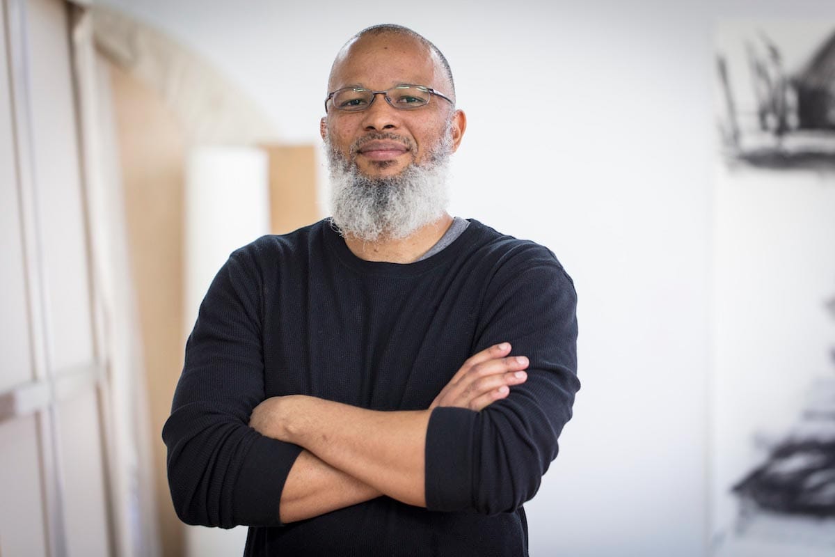 Meleko Mokgosi, pictured from the waist-up, stands with his arms crossed, wearing a black sweater, in his art studio.