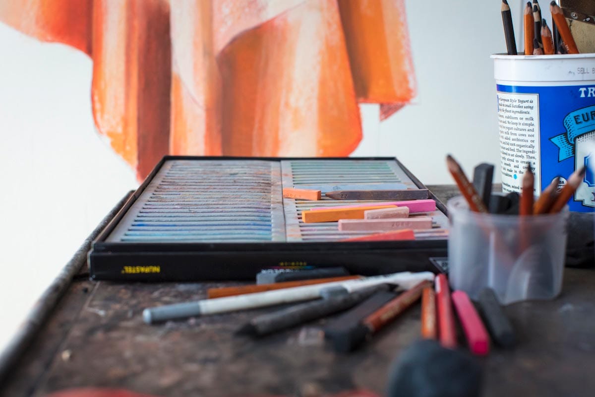 A box of used pastels is pictured on a table with scattered pencils, in front of a section of an orange artwork.