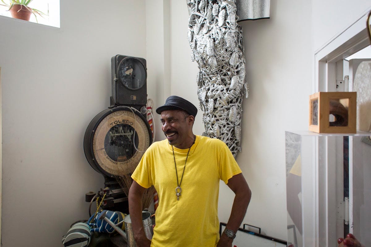 Nari Ward is pictured from the waist-up wearing a yellow shirt, smiling and looking off to the side, with his artworks behind him.