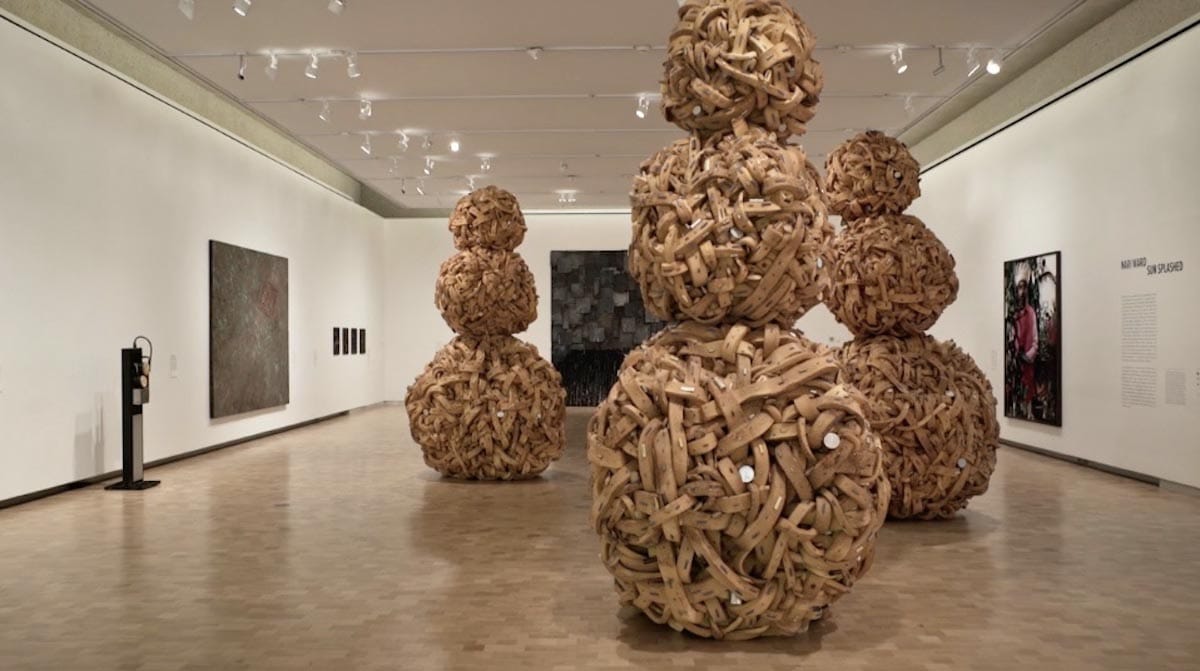 Three large art-pieces each consisting of three large balls stacked atop one another, stand on the floor of a gallery space with other artworks hanging on the walls.