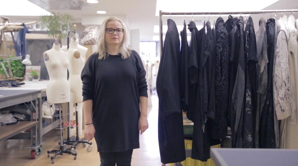 Natallia Pilipenka is pictured standing in a work space alongside a clothing rack and hanging garments, with mannequins and fabric behind her.