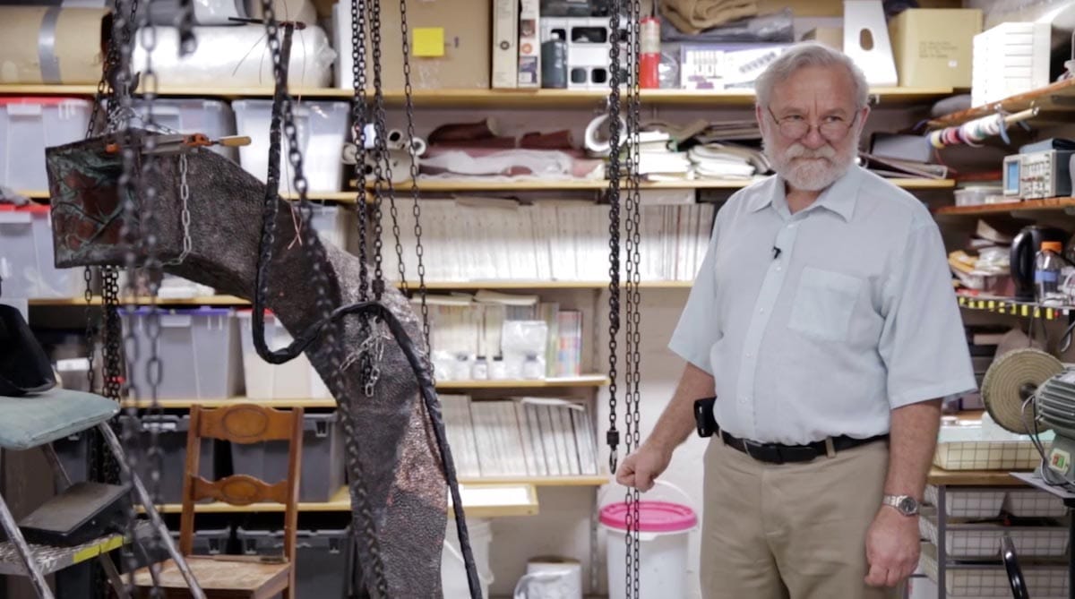 Peter Walter is pictured standing in a workshop with shelves filled with paper, books, and tools behind him.